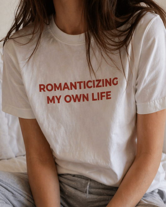 Person wearing a white t-shirt with red text 'ROMANTICIZING MY OWN LIFE' sitting on a bed.