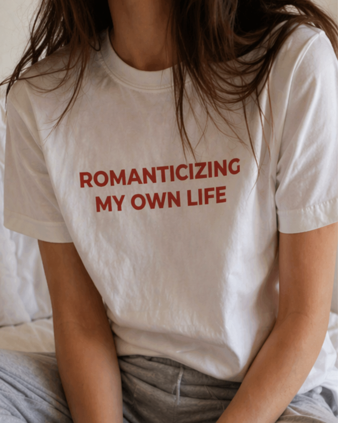 Person wearing a white t-shirt with red text 'ROMANTICIZING MY OWN LIFE' sitting on a bed.
